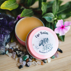 Eye balm in pink tin with "Eye Balm" text, Ninas Bees brand, surrounded by flowers