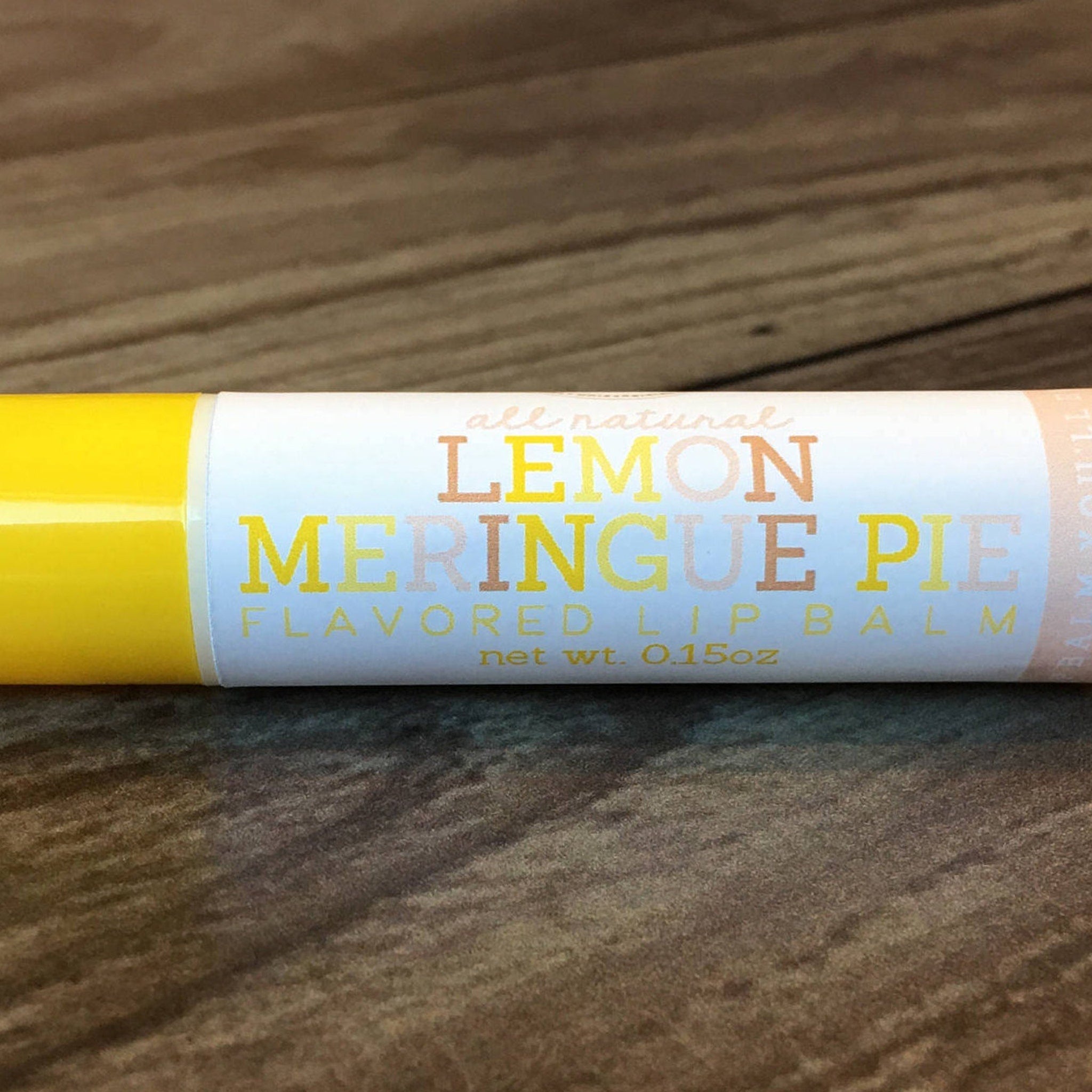 Lip balm tube with yellow and white, "Lemon Meringue Pie" text, on wood grain background