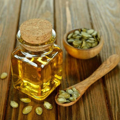 Pumpkin seed oil in a clear glass bottle with a cork stopper, wooden spoon, and seeds on a wooden table