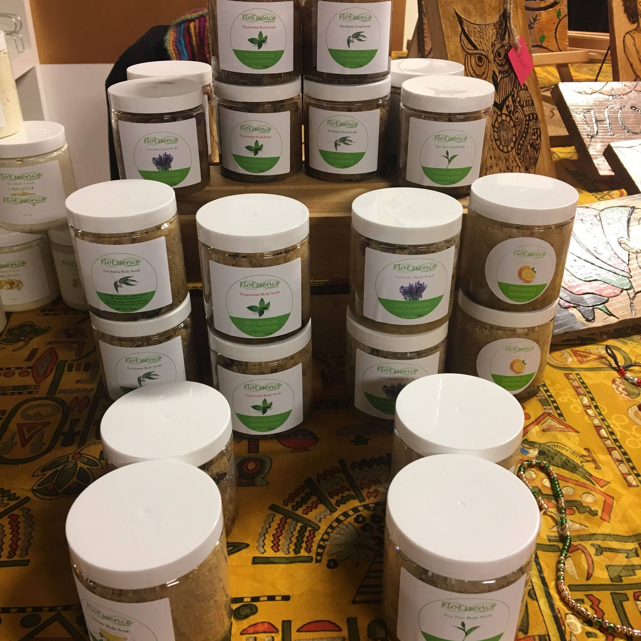 Facial scrub jars, clear plastic with white lids, FloEssence labels, on a yellow patterned surface