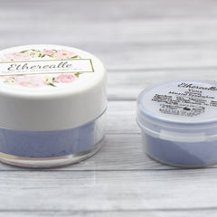 Eyeshadow pots with white lids, blue powder, and Etherealle branding on a wooden surface