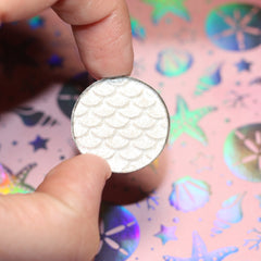 Eyeshadow single in white with scale pattern, held by fingers, on pink background