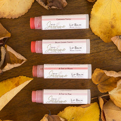 Lip balm tubes with pink, red, and white labels, Wild Jasmine brand, on a wooden surface with leaves