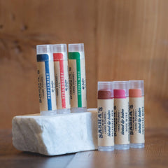 Lip balm tubes with blue, red, and green labels, on white stone, Sasha's brand visible