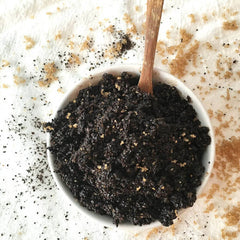 Body scrub in white bowl with wooden spoon, dark brown mixture with brown and tan granules scattered around