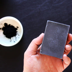 Soap bar held in hand, gray rectangular shape, with bowl of black powder in background
