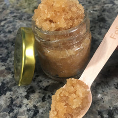 Sugar scrub in a glass jar with a wooden spoon, light brown granules, gold lid, and wooden spoon