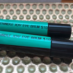 Eye balm tubes, teal and black, with "REKINDLE JOY" text, on textured surface
