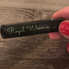 Lip balm tube with black body, green "Royal Winnie" text, held by hand with red nails