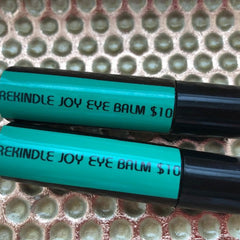 Eye balm tubes, teal and black, with "REKINDLE JOY EYE BALM" text visible, on textured surface