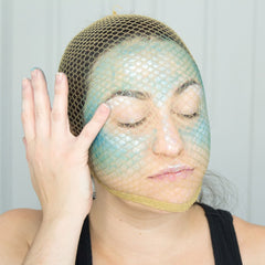 Face paint with blue and white scales, covered with beige net, hand touching face