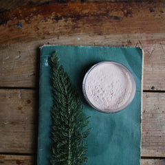 Powder in clear bowl with pinkish hue on teal book and wooden surface with green sprig