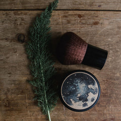 Makeup brush with brown bristles, black handle, and round compact with abstract design on wood surface