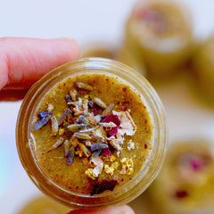 Topical salve with green, brown, and purple floral elements in a clear glass jar, held by a hand