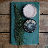 Finishing powder in clear jar with light pink powder on teal book with green sprig on wood table
