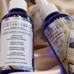 Floral water bottles, blue glass with white sprayers, FLEURESSENCE label visible, on white fabric surface