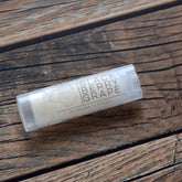 Lip balm tube with white label, clear plastic, and text "Blackberry Grape" on wood background