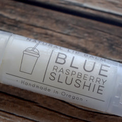 Lip balm tube with white label, gray text "Blue Raspberry Slushie", and a cup graphic on a wooden surface