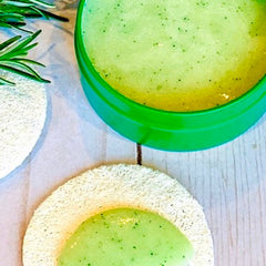 Green scrub with green container, white loofah, and rosemary sprig on a wooden surface