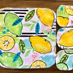 Reusable cloth pads with yellow lemon and green lime fruit pattern, white stitching detail, on wood surface