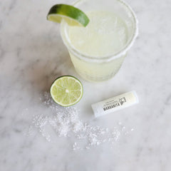 Lip balm tube with white packaging, "Margarita" text, and lime slice on marble surface