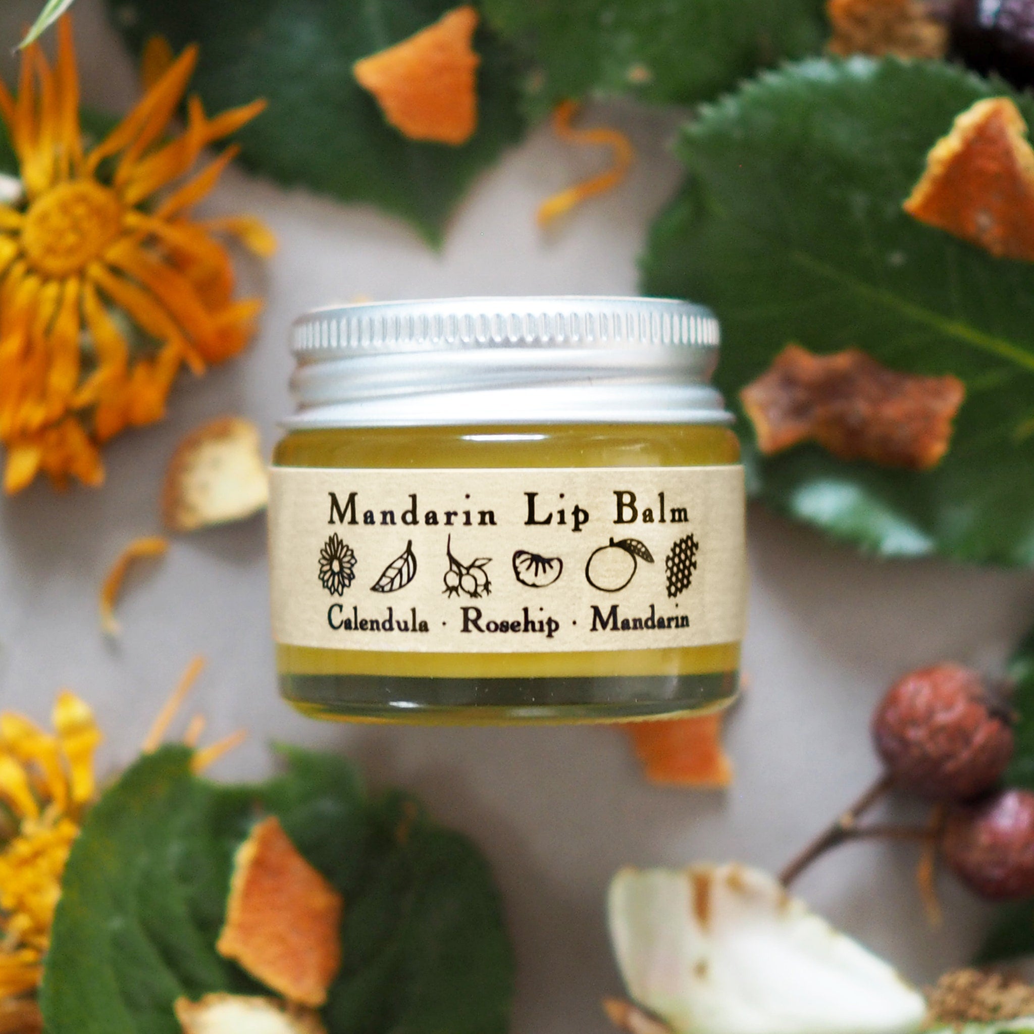 Mandarin lip balm jar with silver lid and label, surrounded by orange and green foliage