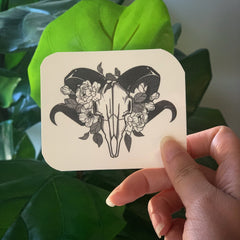Sticker with black and gray ram skull design, floral pattern, held by hand