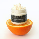 Facial scrub with white cream, glass jar, and orange slice, Bellaroma Boutique label visible