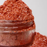 Eye shadow glitter in pink copper color, in a clear jar with product spilling out