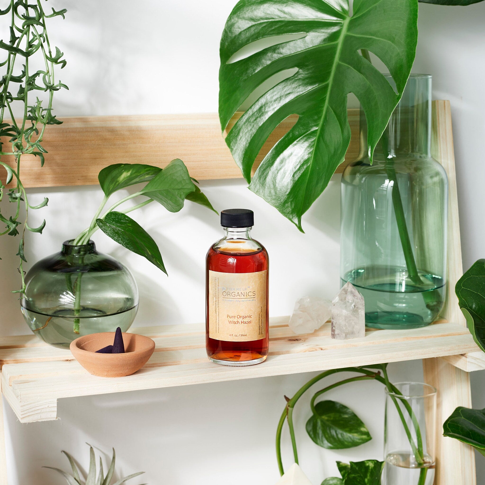 Organic Witch Hazel Toner, amber liquid in clear glass bottle, Butter Me Up Organics label visible on shelf with plants