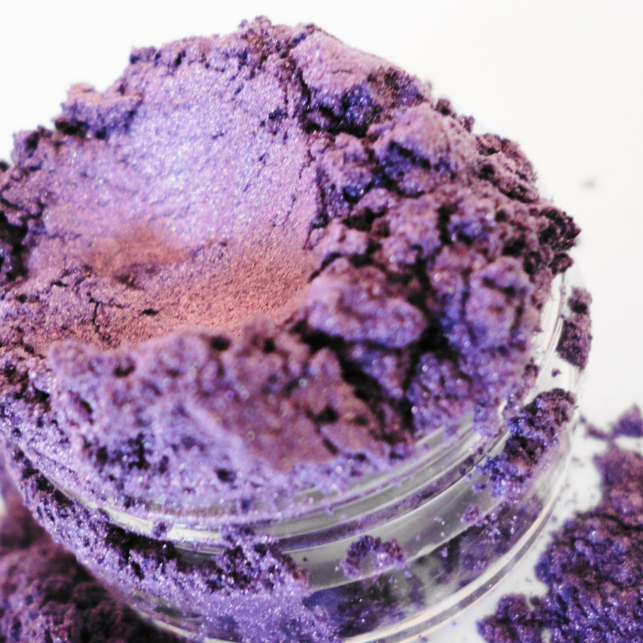 Eyeshadow powder in purple shimmer, visible in a clear glass jar, on a white background.