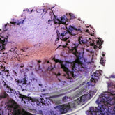 Eyeshadow powder in purple shimmer, visible in a clear glass jar, on a white background.
