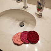 Face Scrubbies, three crochet rounds in pink, fuchsia, and red, near a sink