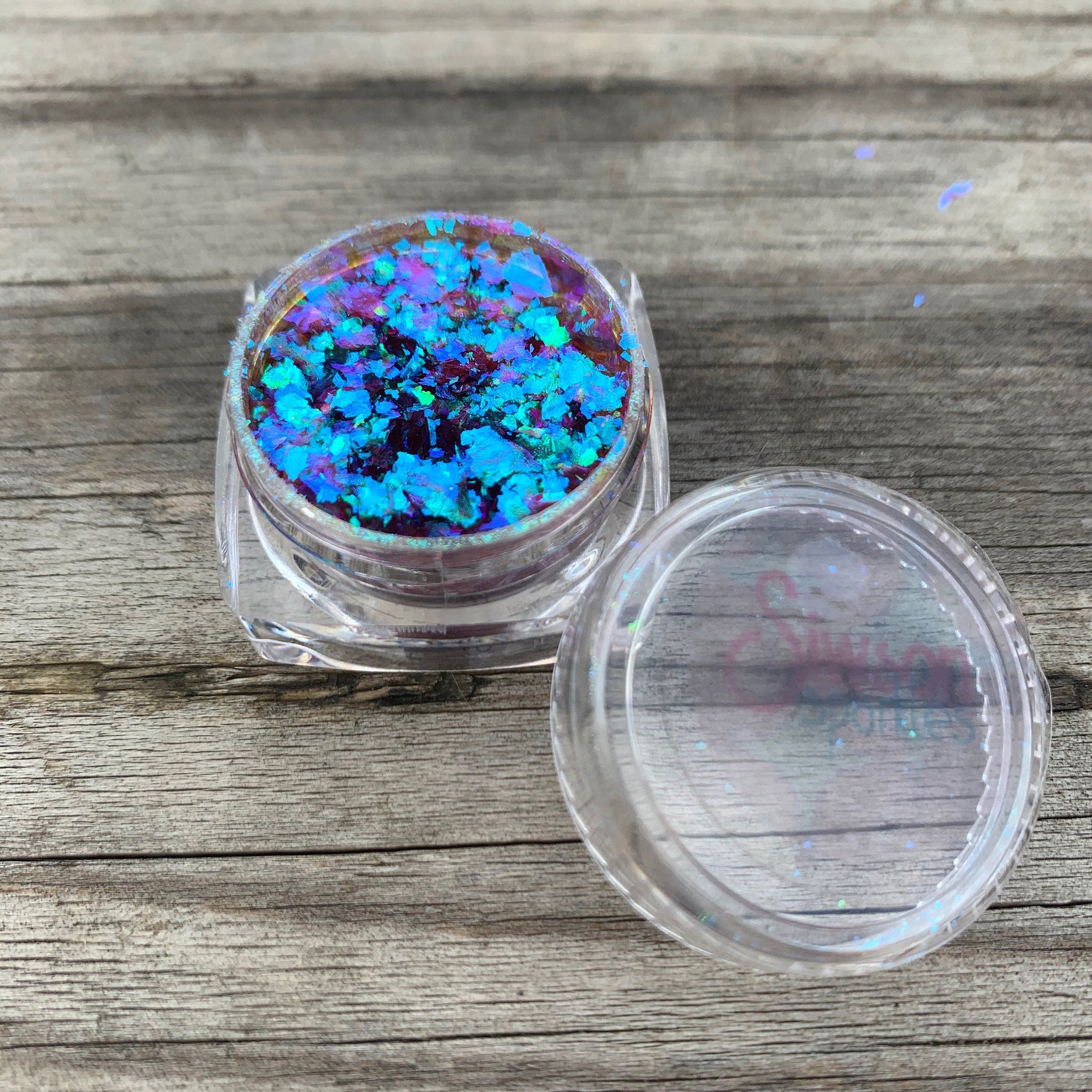 Close-up of VENUS multichrome eye shadow flakes shimmering with shifting iridescent hues in a sleek black container