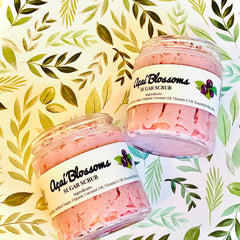 Sugar scrub jars with pink scrub, clear glass, and "Açaí Blossoms" label on green leaf background.