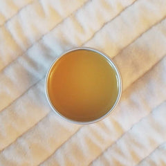 Lip balm in a silver tin with golden yellow color, sitting on white quilted fabric