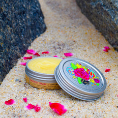 Lip balm in silver tin with floral lid design and yellow balm, on sand with rose petals