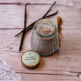 Sugar scrub in glass jar with wooden spoon and vanilla beans on wood surface, DalaNaturalBeauty label