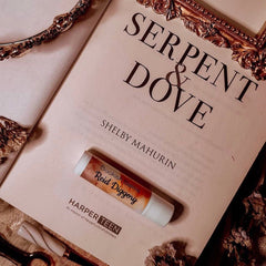 Lip balm tube with white and orange label, resting on a book, Serpent & Dove brand
