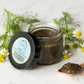 Facial cleanser jar with black lid, clear glass, and dark brown scrub inside