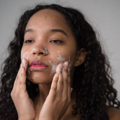 Facial cleanser application, woman with dark curly hair and white foam on face, hands touching skin