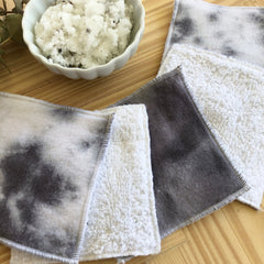 Washcloths with tie-dye pattern, white terrycloth, grey trim, and a bowl of scrub on wooden surface