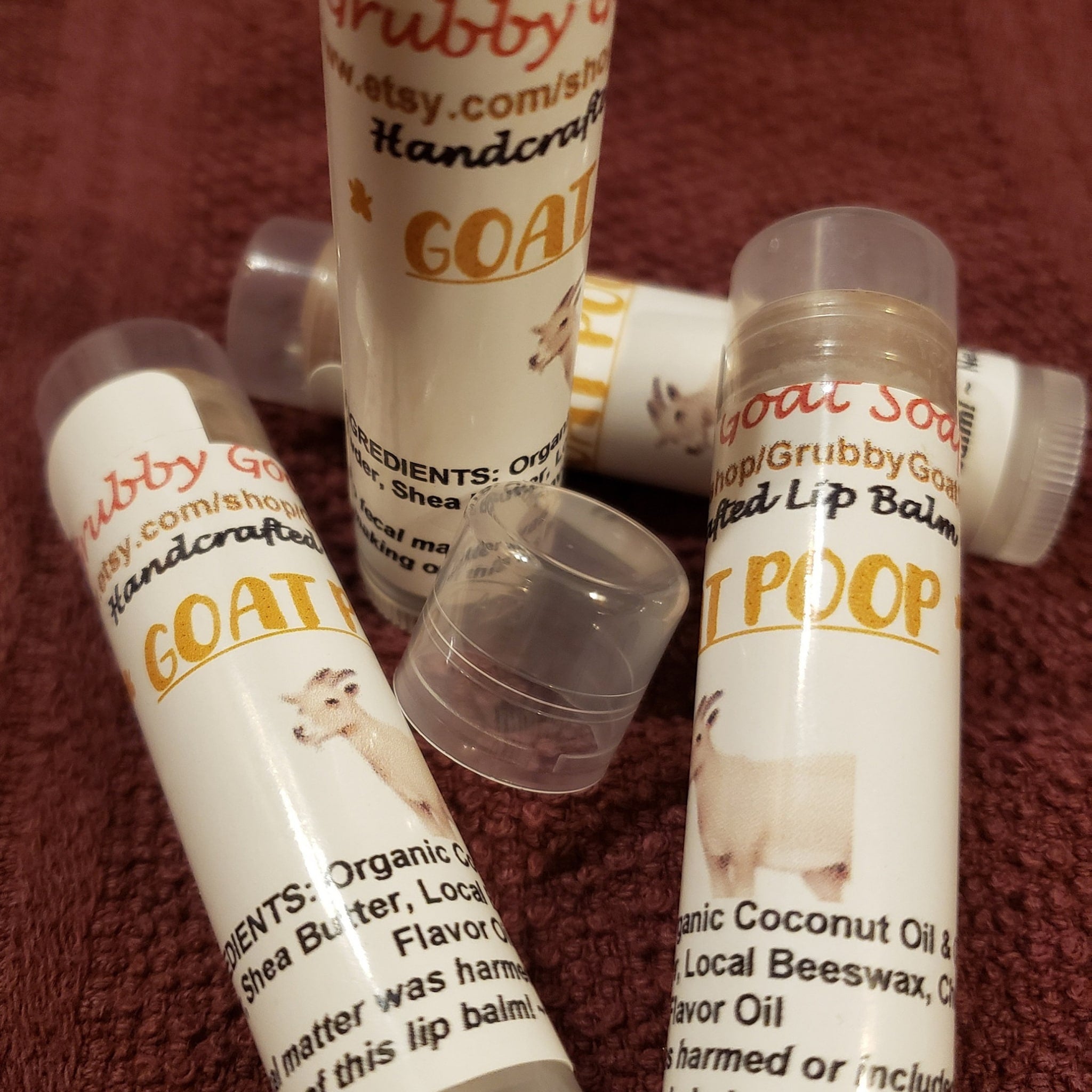 Lip balms white tubes with goat graphic and GrubbyGoatSoap text, clear cap on red fabric