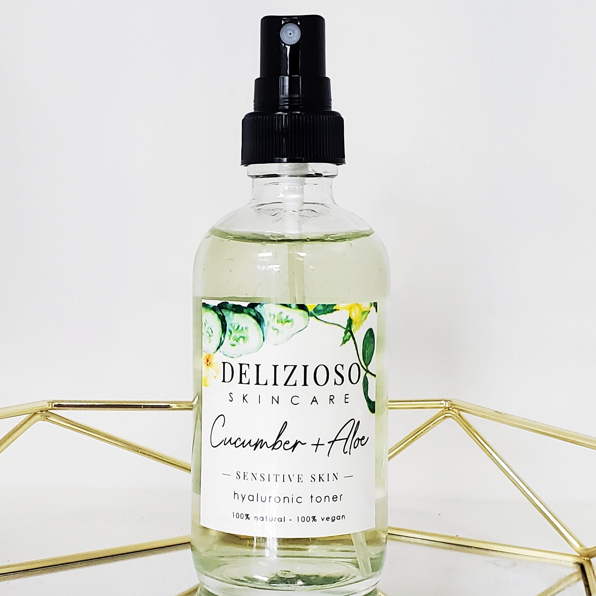 Cucumber toner in clear glass bottle with black spray top, Delizioso Skincare label, and gold geometric stand