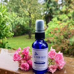 Face tonic bottle with blue glass, black cap, and "LE NOIR BLEU" label, with pink roses on wood surface