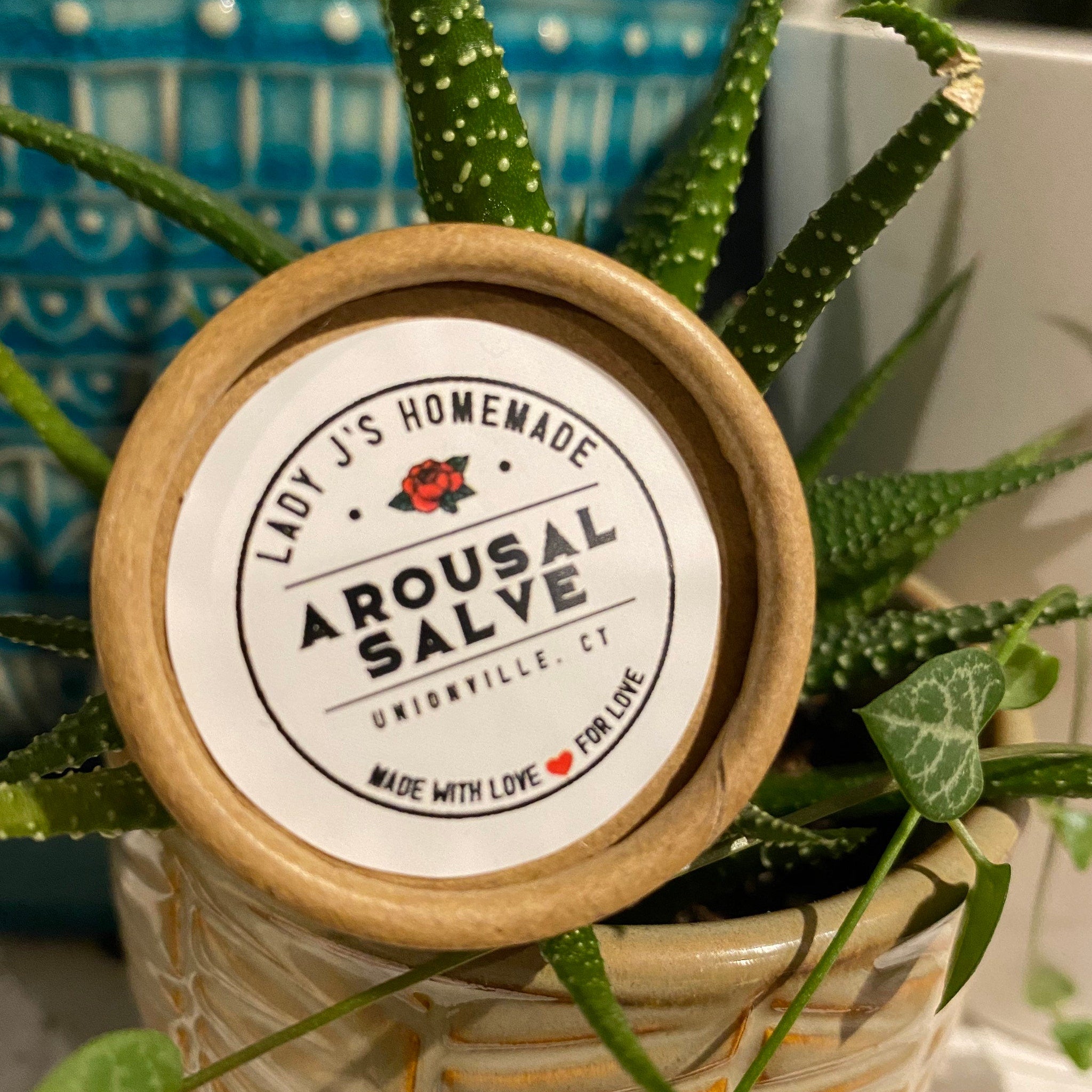Personal Lubricant container, brown cardboard, white label with Lady J's Arousal Salve text, green plant in background