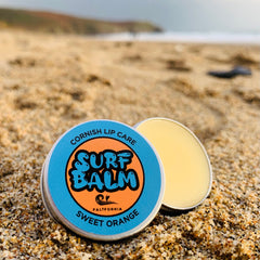 Surf balm tin with blue and orange label, silver metal container, and yellow balm on sand
