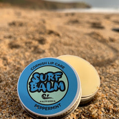 Surf balm tin with blue and green label, silver metal, and yellow balm on sand