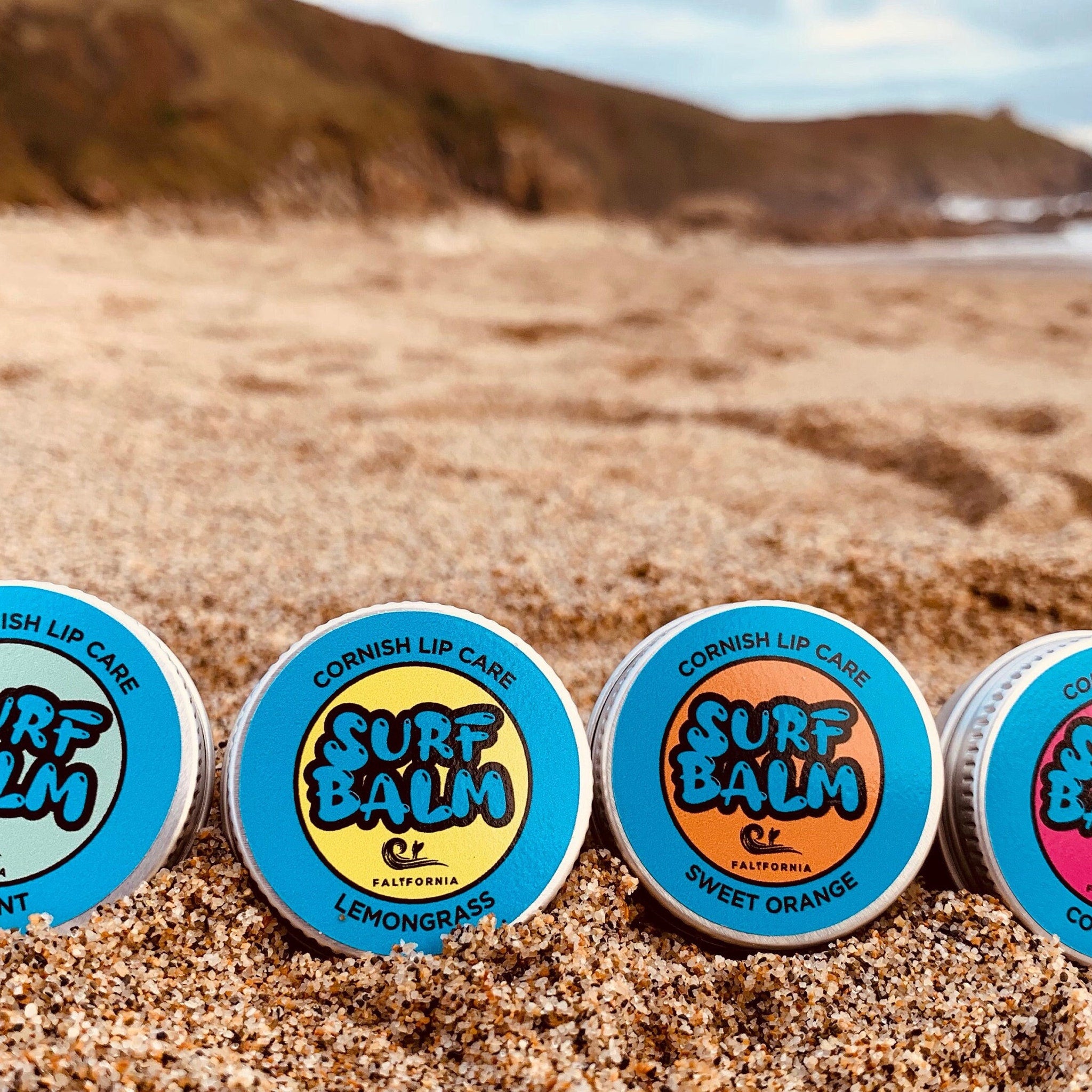 Lip balms with blue, orange, and white lids, SURF BALM text, on sand
