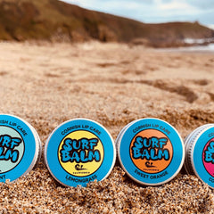 Lip balms with blue, orange, and white lids, SURF BALM text, on sand